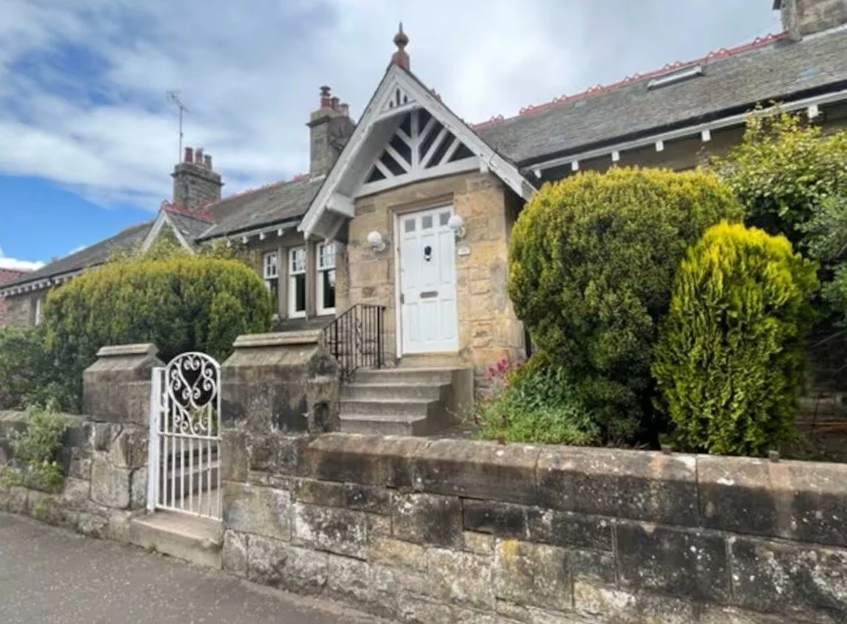 A historic house with stone construction, a white front door, small steps leading up to it, a stone fence, and lush green bushes in front.