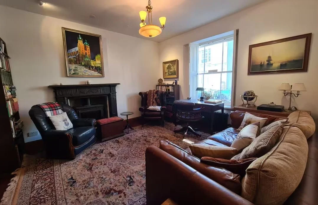 A living room with a bookshelf, two armchairs, a sofa, a desk by the window, a fireplace, framed art, and a decorative rug.