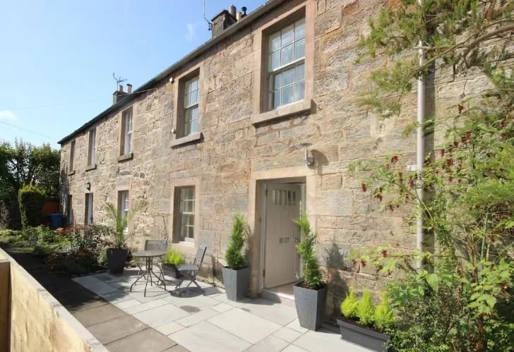 Exterior view of a stone house with a small patio featuring potted plants, outdoor furniture, and a stone pathway.