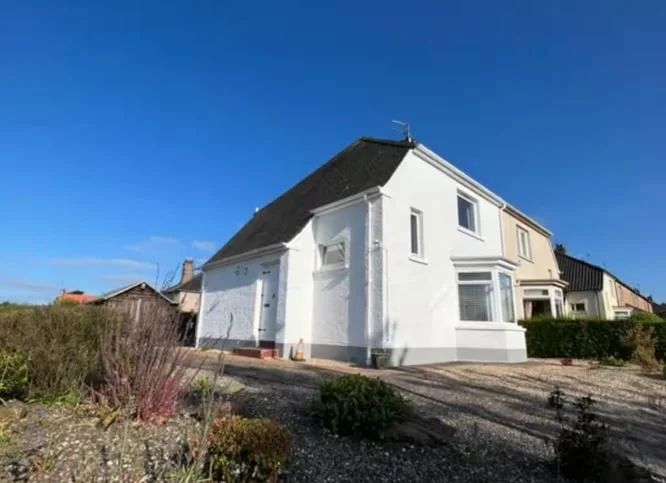 A white, two-story house with a black roof under a clear blue sky, surrounded by a garden with shrubs and gravel driveway.