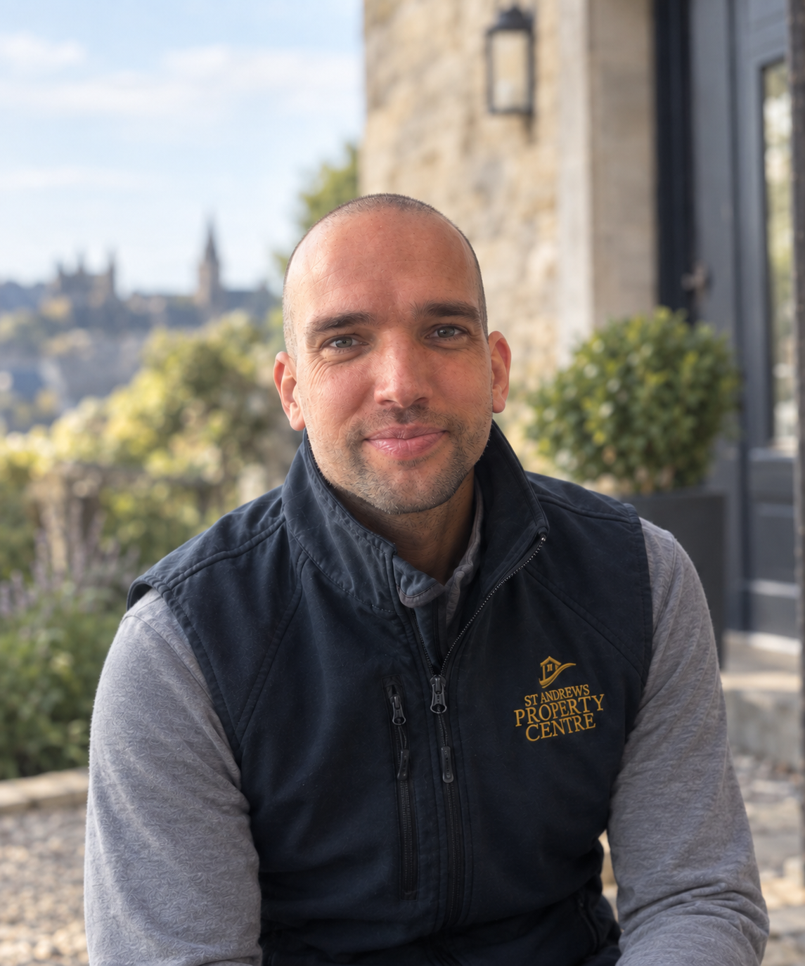 A man with short hair and a slight beard sits outdoors on a sunny day, wearing a black vest with 'St Andrews Property Centre' logo, with a blurred historic stone building and cityscape in the background.