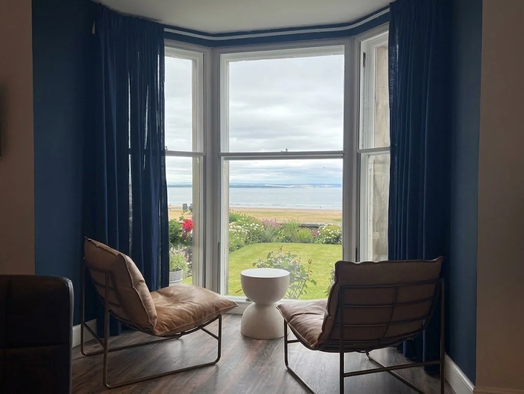 A cozy sitting area with two brown leather chairs facing a large window with blue curtains, overlooking a garden and a body of water.