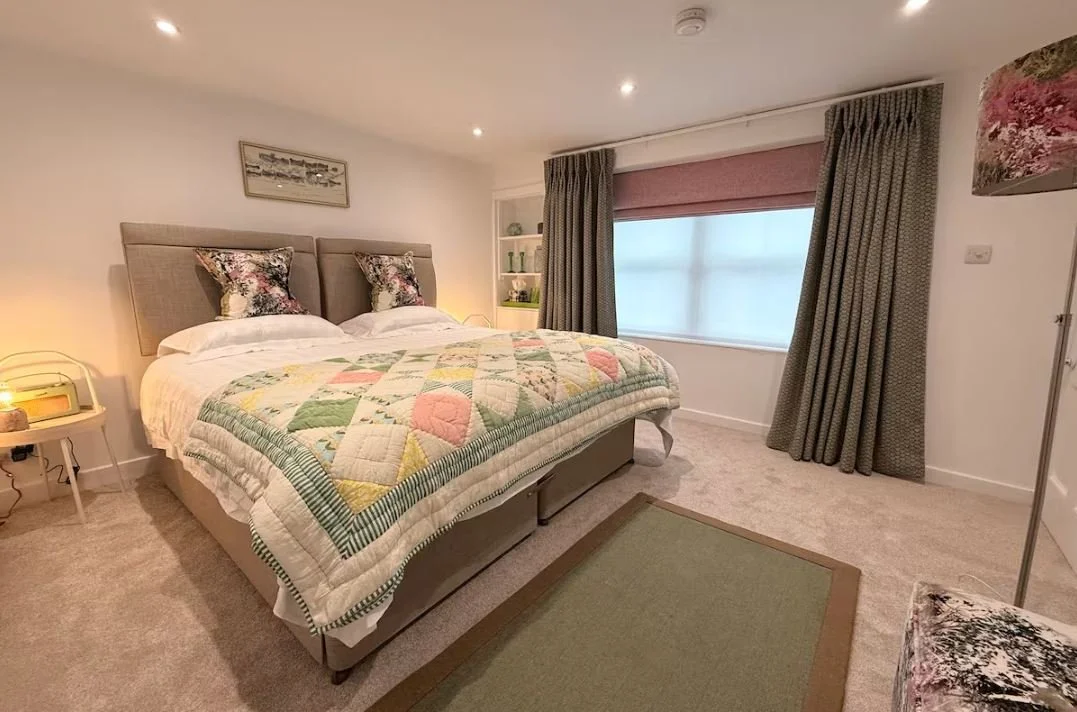 A cozy bedroom with a large bed with a colorful quilt and floral pillows, bedside table with a lamp, a window with curtains, and a carpet on the beige carpeted floor.