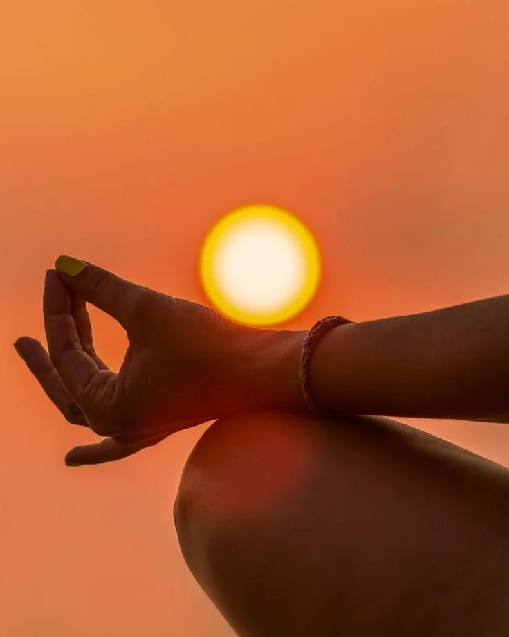 A person sitting with their legs crossed and hand extended, with the setting sun in the background and the palm facing outward against a vibrant orange sky.