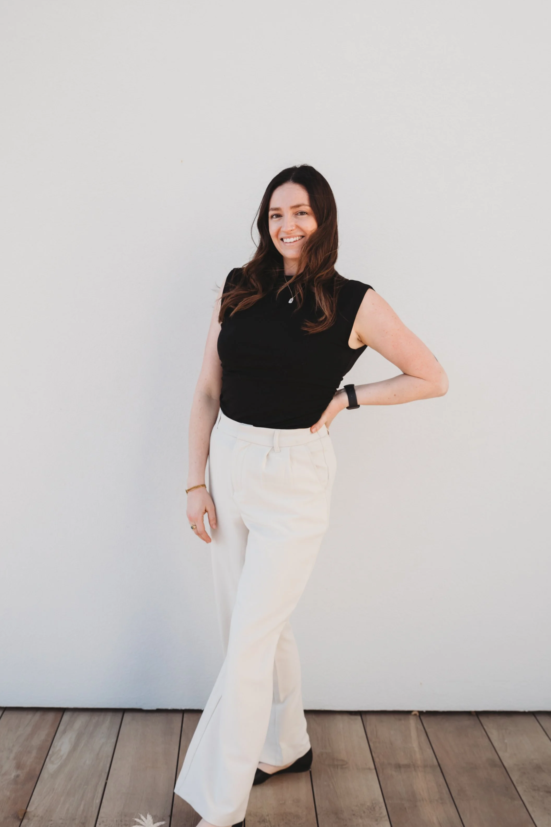 A woman standing against a plain white wall, smiling, with hand on her hip, wearing a sleeveless black top and high-waisted white pants.