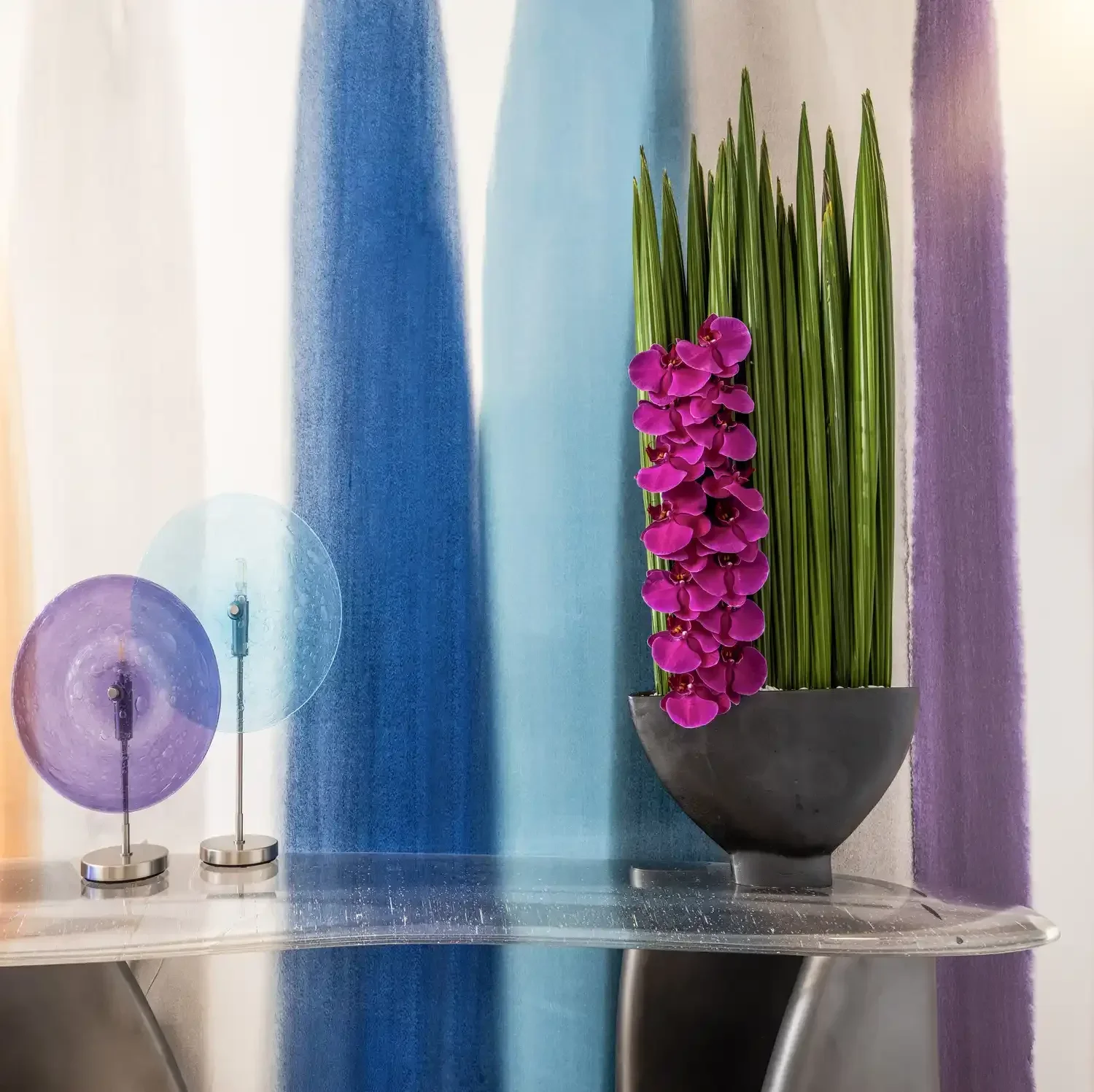 A potted plant with pink orchids and tall green grass-like leaves on a glass table, flanked by two modern purple and blue glass table lamps with metal bases, against a colorful abstract wall background.