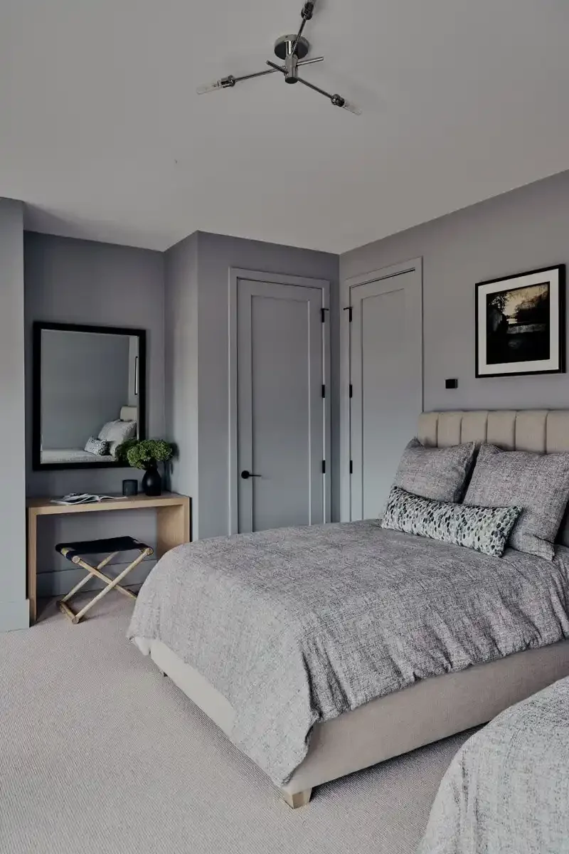 A bedroom with a bed, a mirror, a small vanity table, and a framed picture on the wall. The room has light gray walls and a beige carpet.