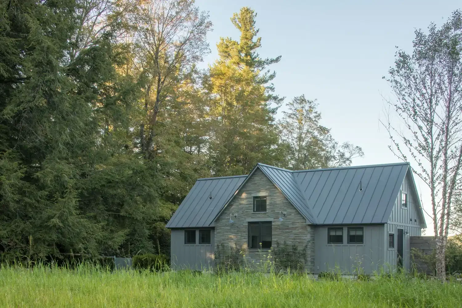 A modern two-story house with gray siding and a gabled metal roof, surrounded by green trees and grass, in a rural setting during daytime.