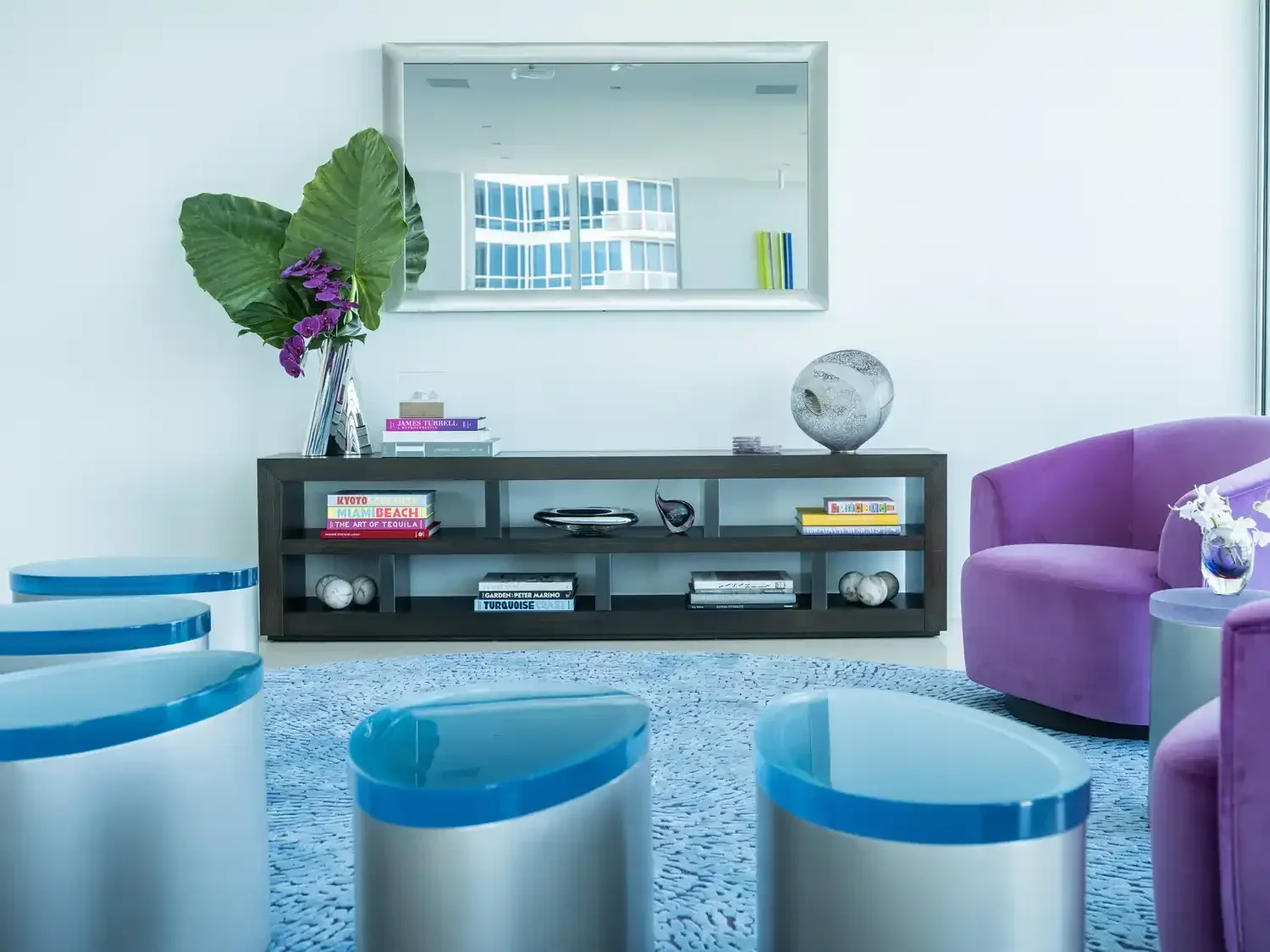 Modern living room with purple armchairs, a black console table with books and decorative objects, a large mirror, and a tall plant with large green leaves and purple flowers.