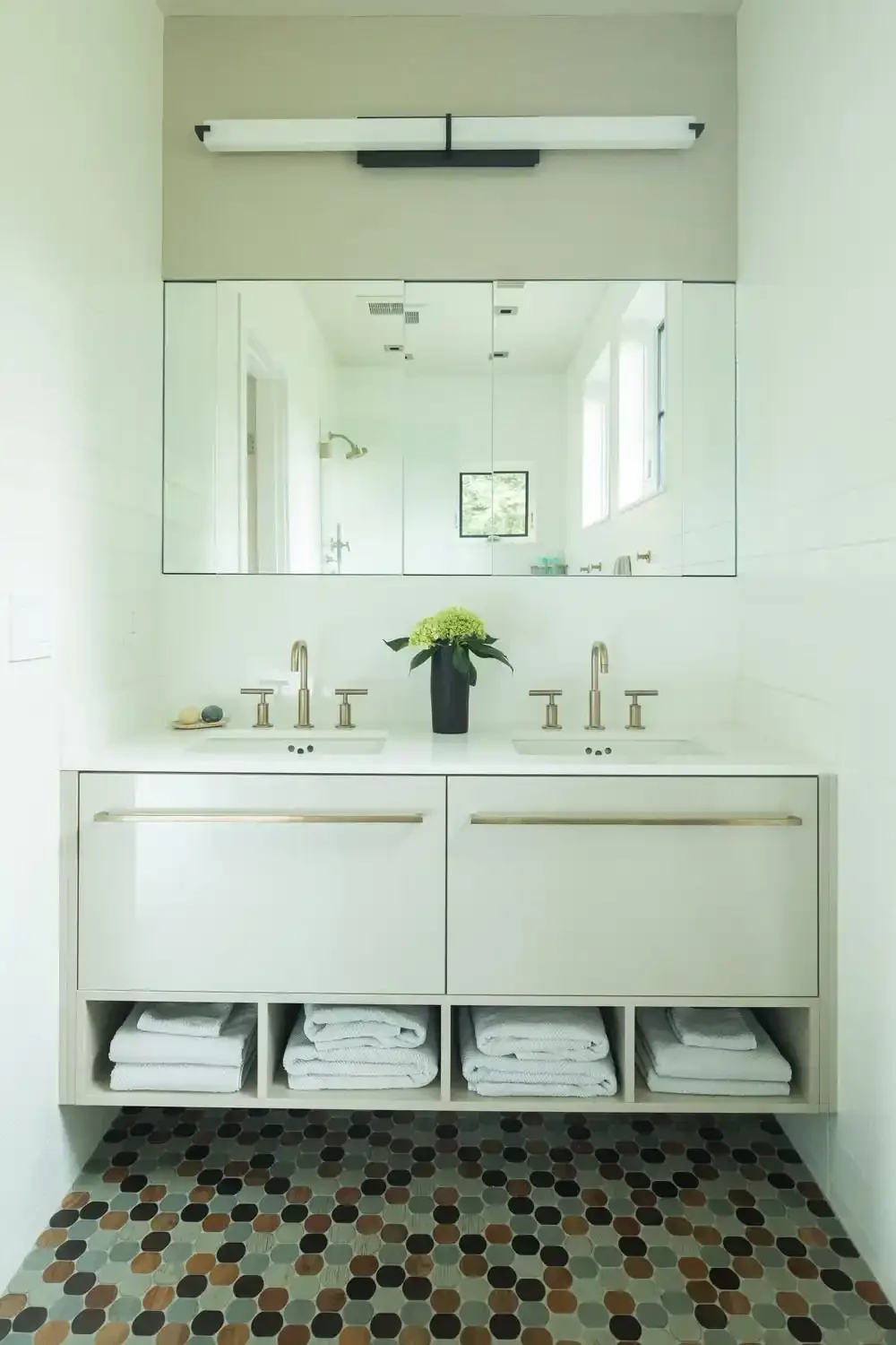Modern bathroom with double sink vanity, large mirror, flower vase, towels, and hexagonal tile floor.
