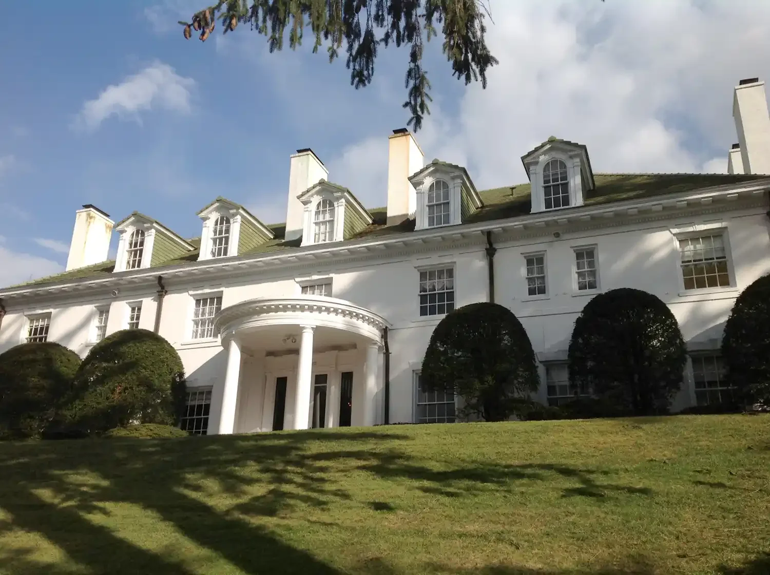 A grand white mansion with multiple dormer windows, surrounded by manicured trees and grass, under a partly cloudy sky.