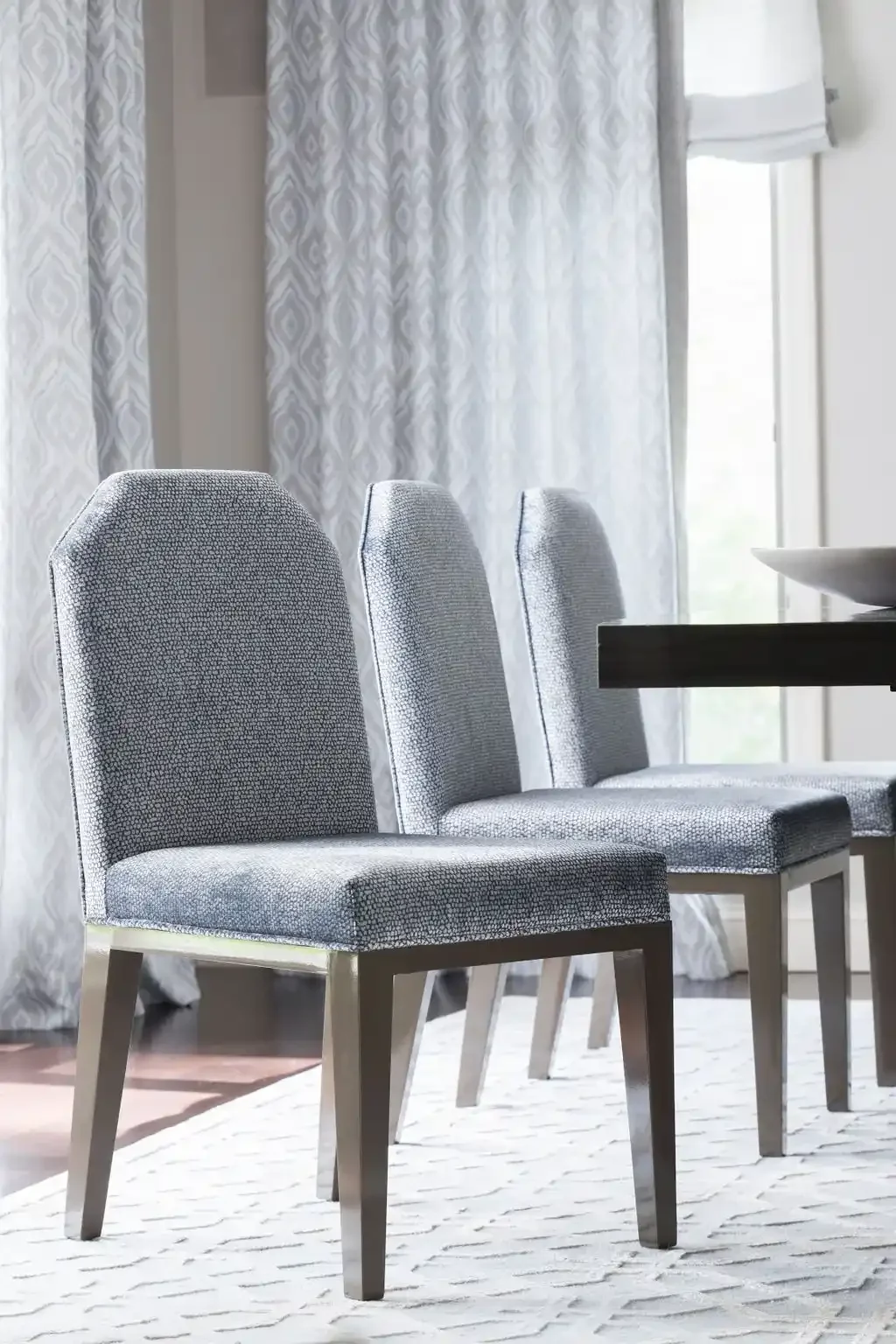 Three upholstered dining chairs with gray fabric and wooden legs arranged around a dark wooden table in a bright room with white patterned curtains.