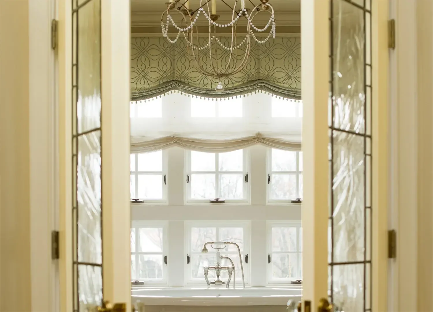A view of a bright bathroom seen through an open glass door with a decorative frame. The bathroom features a large window with curtains, a chandelier hanging from the ceiling, and a bathtub with a vintage faucet.