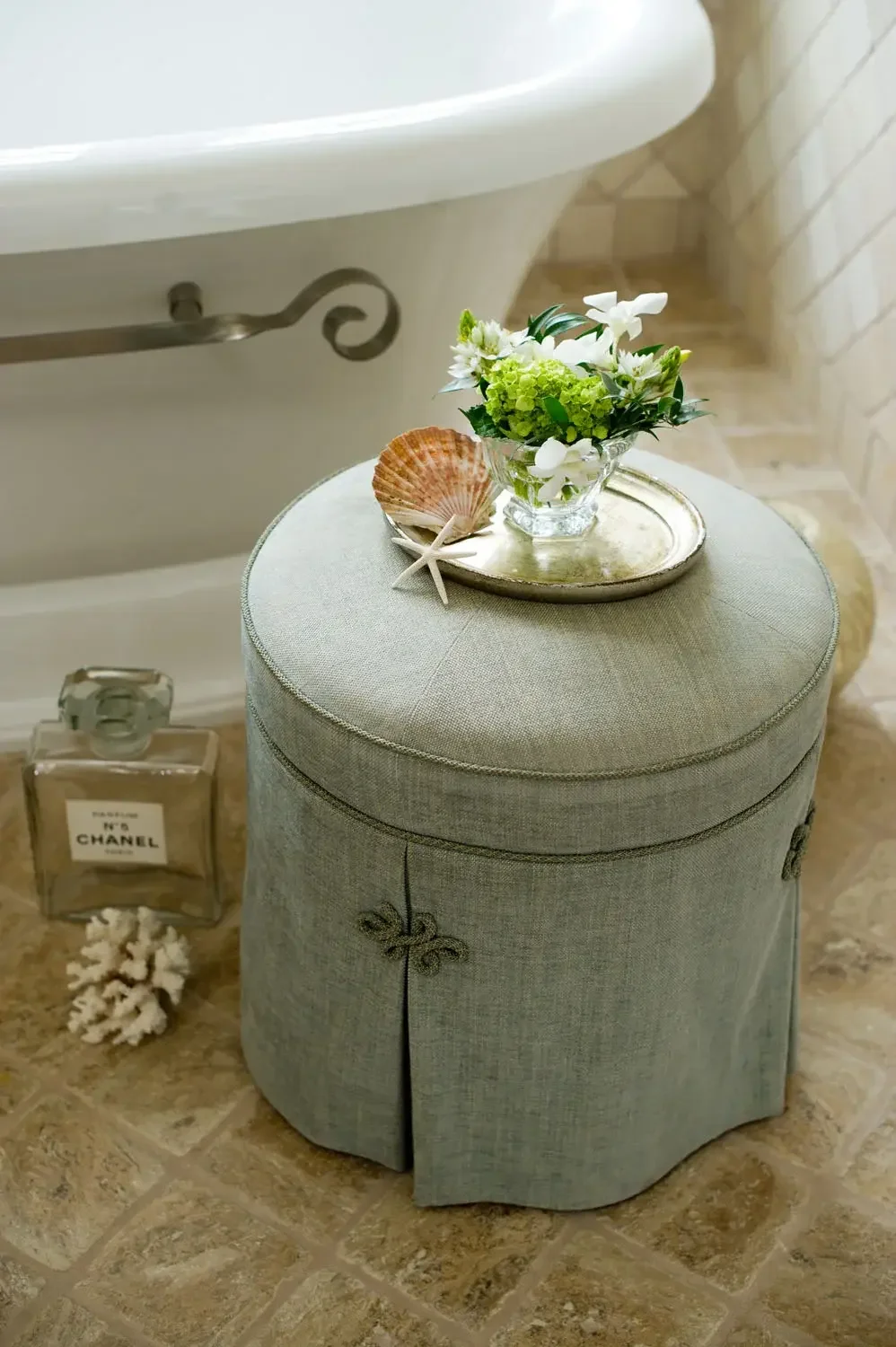 A small round upholstered ottoman with a grey fabric cover, topped with a glass bowl of white and green flowers and a silver tray. Next to the ottoman on the floor is a bottle of Chanel No. 5 perfume and a seashell with a starfish, near a bathtub wit