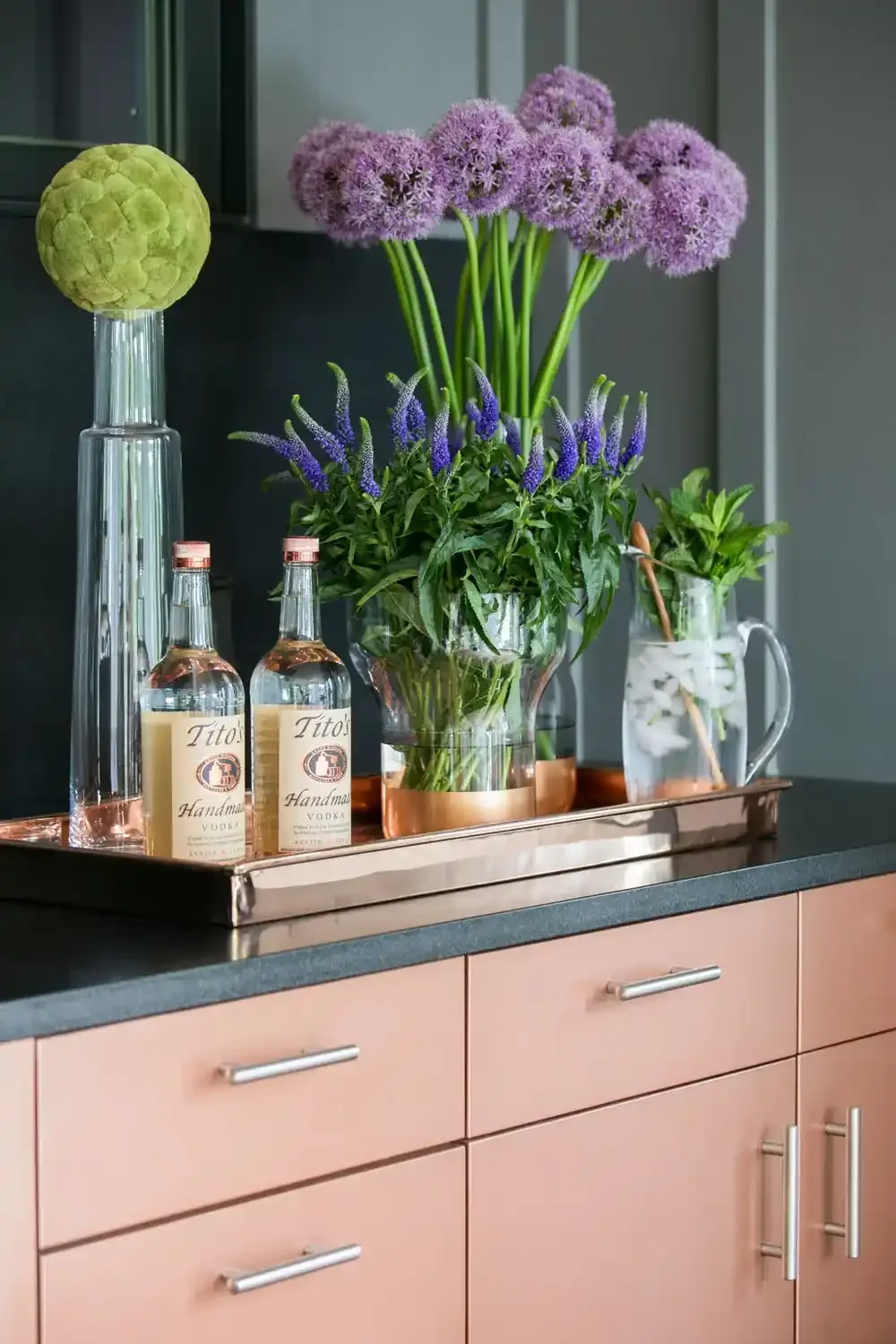 Vase with purple and lavender flowers, two bottles of Tito's handmade vodka, and a glass pitcher with greenery on a pink cabinet.