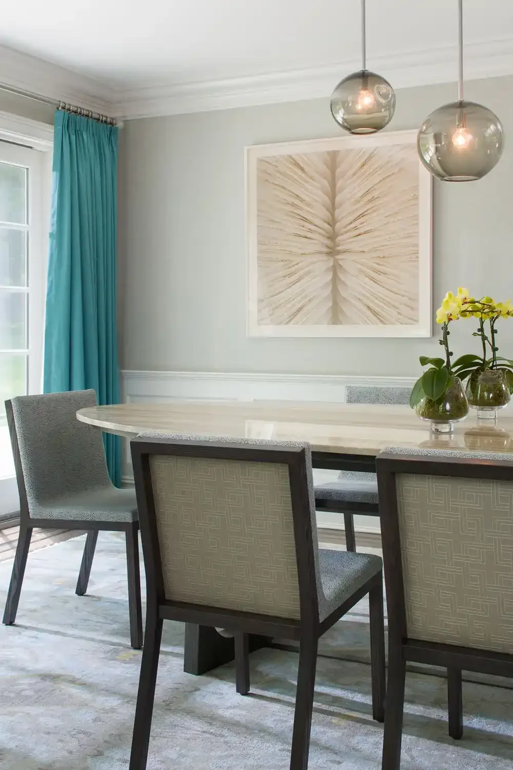 A dining room with a white oval table, four chairs with patterned beige upholstery, a large abstract wall art, a vase with yellow orchids, and a window with turquoise curtains, modern spherical hanging light fixtures.