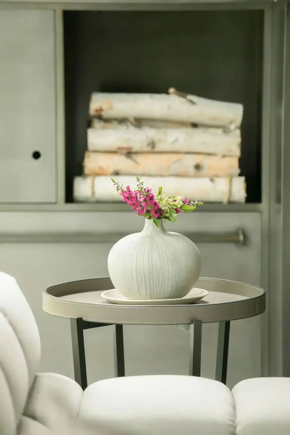 A white round vase with pink and white flowers on a small round table, in front of a cabinet with stacked logs.