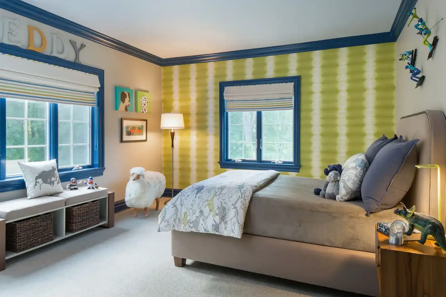 A bedroom with two windows, green striped wallpaper, a beige bed with gray pillows and stuffed animals, a white bench with pillows and toy robots, a lamp, artwork on the wall, and colorful wall decals on the ceiling.