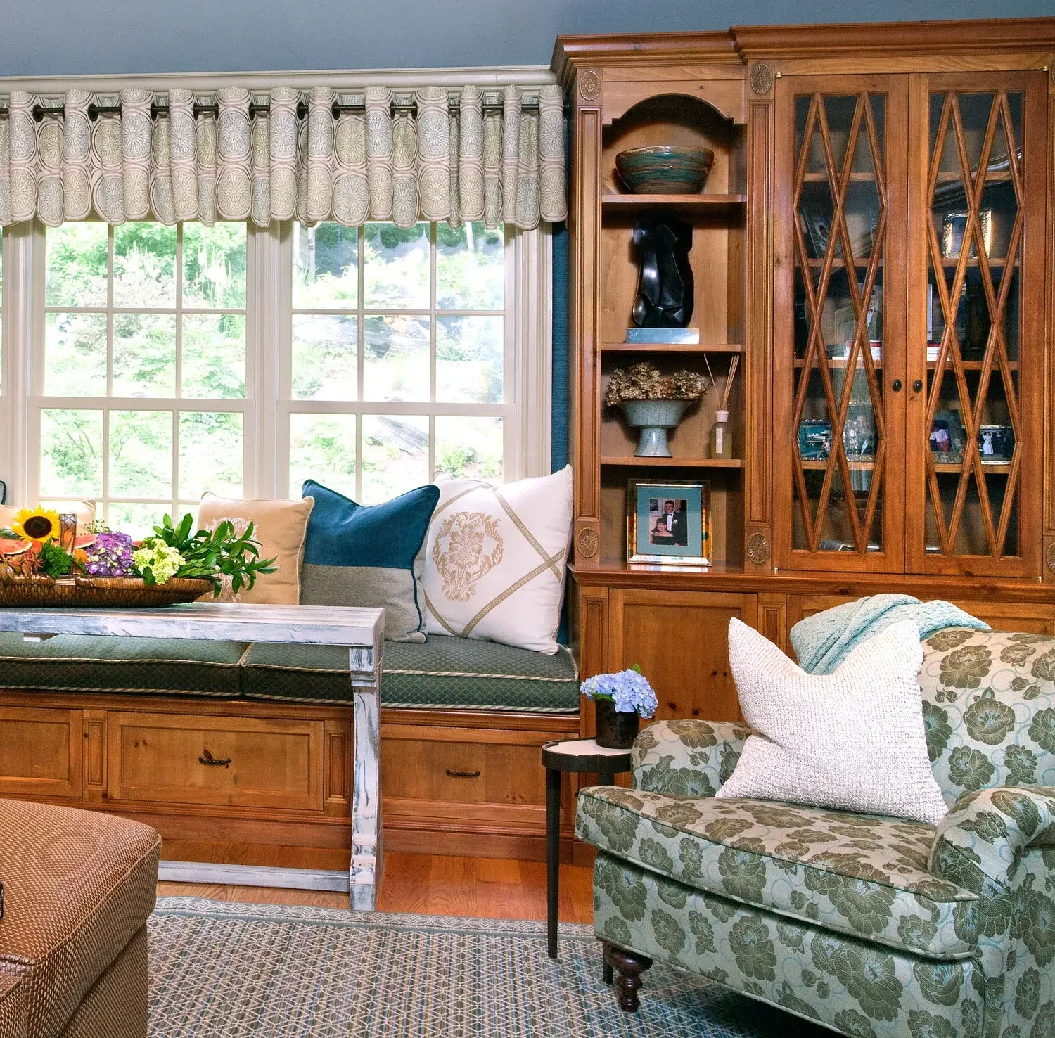 Living room with window seat, floral and solid pillows, wooden cabinet with glass doors, and armchair with floral pattern and pillows.