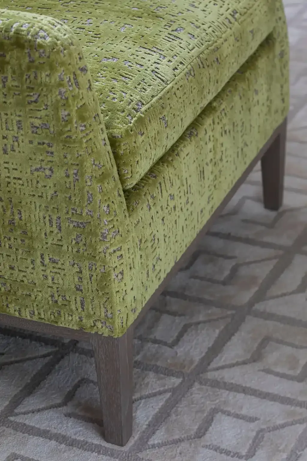 Close-up of a green upholstered armrest on a sofa with textured fabric, featuring a pattern of geometric lines, and wooden legs, on a patterned rug floor.