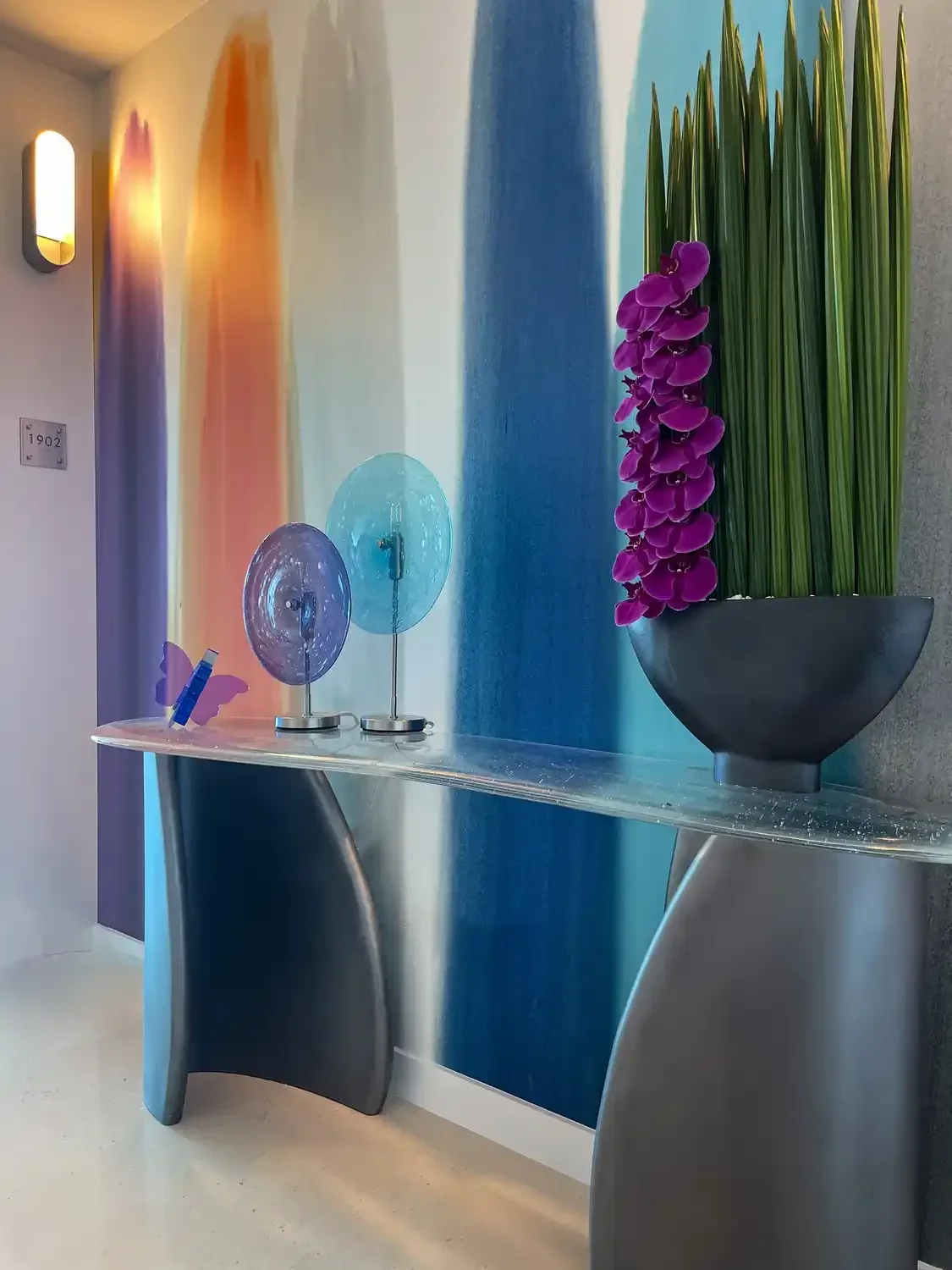 Decorative hallway with colorful wall art, purple and blue glass objects on a sleek metal and glass table, black bowl with tall green grass and purple flowers.