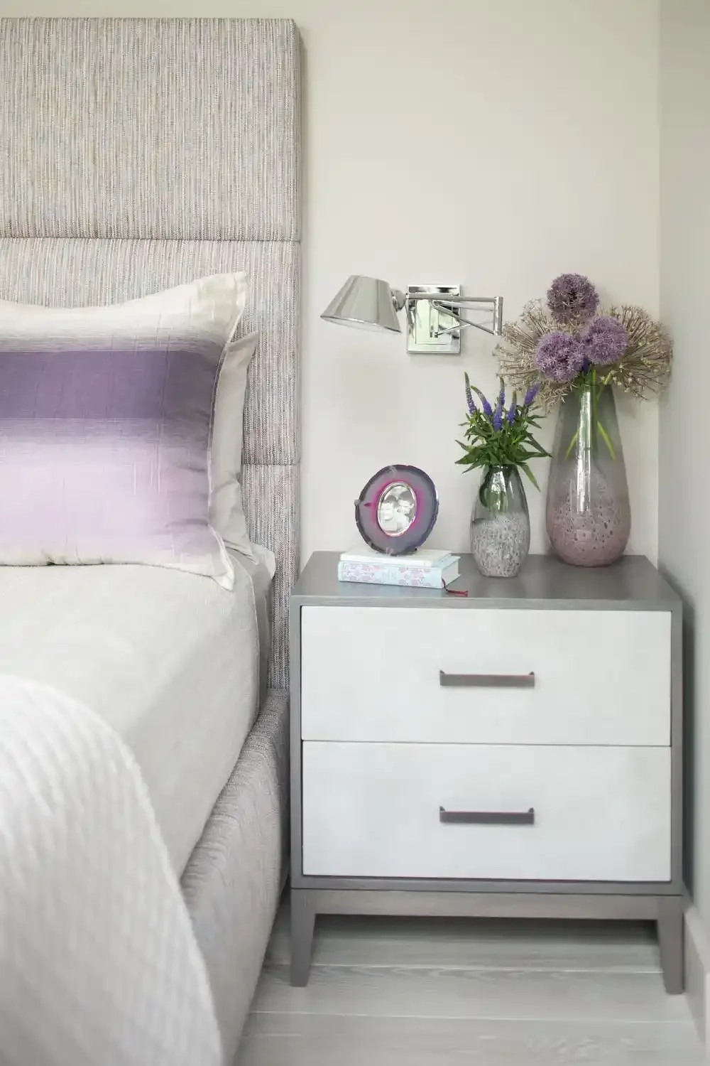 A bedside table with two drawers, topped with decorative vases filled with purple flowers and a small clock, situated next to a bed with a tufted upholstered headboard and a purple and white bedding set, against a white wall.
