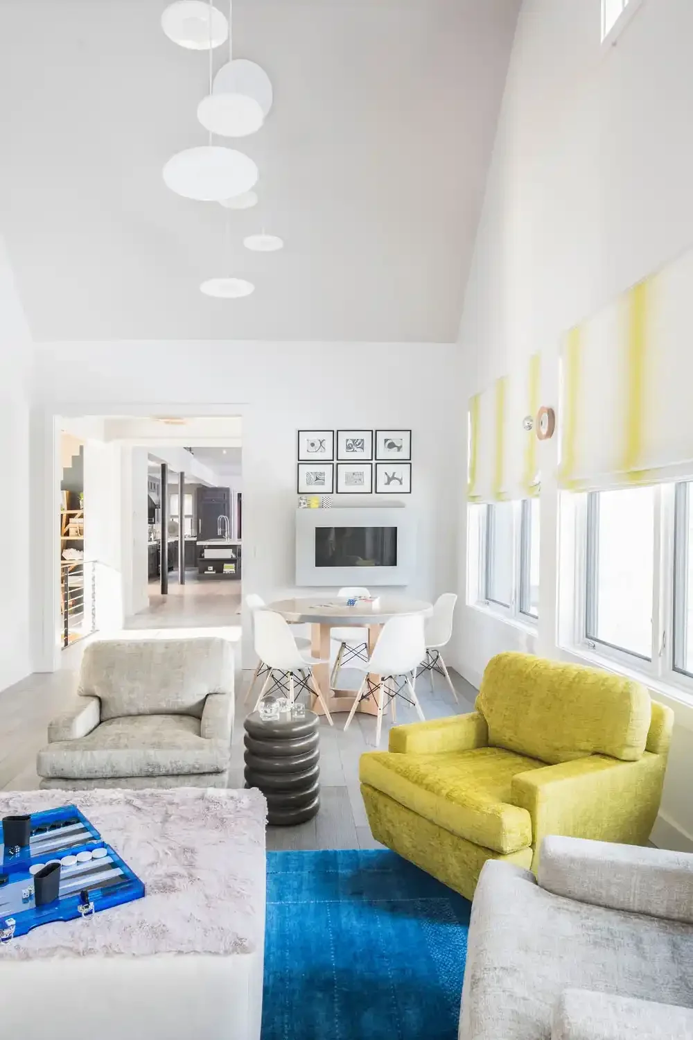 Bright living room with white walls, yellow armchair, beige armchair, blue rug, contemporary furniture, multiple windows with yellow blinds, round dining table with white chairs, flat-screen TV, and modern black and white artwork on wall.