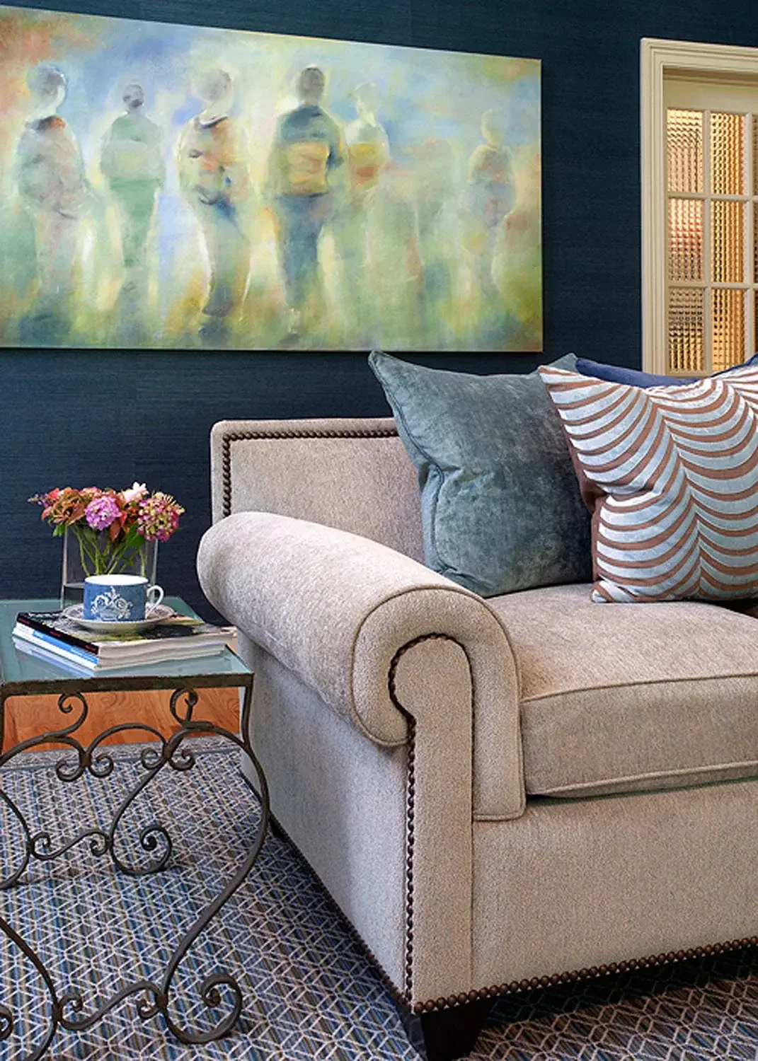 Living room with a beige upholstered sofa, decorative pillows, a glass coffee table with a cup, books, and a vase of pink flowers. A large abstract painting of people hangs on a blue wall behind the sofa. There is a patterned rug and part of a window