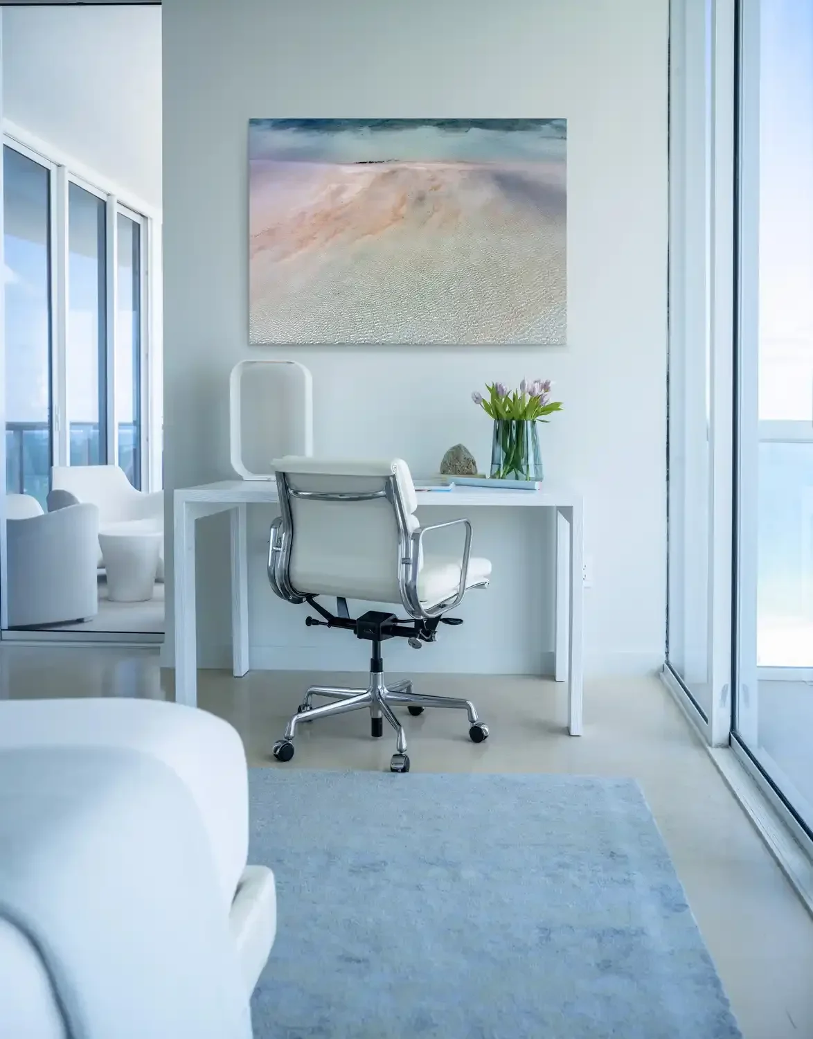 Modern white office with glass walls, a white desk, a white ergonomic chair, a vase with pink tulips, a rock, and a large landscape painting on the wall.