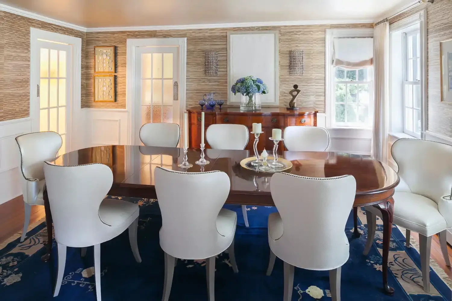 Elegant dining room with a large wooden table surrounded by six white upholstered chairs, candles and glassware on the table, a blue area rug underneath, wood-paneled walls, and large windows with white curtains, a sideboard with blue vases, and deco