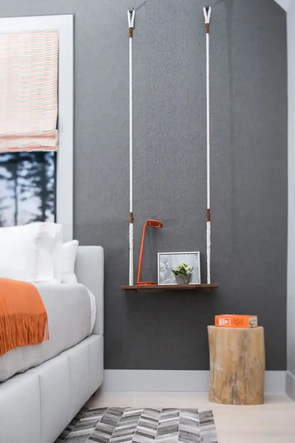 Interior of a bedroom with a white bed, orange throw blanket, a floating wooden shelf with a small plant, a framed mirror, and an orange desk lamp. The wall behind has large climbing ropes hanging from hooks, and a small wooden stool with an orange b