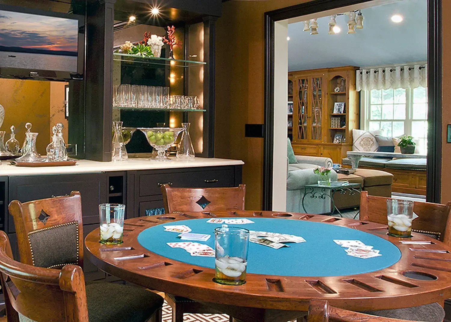 A round wooden poker table with playing cards, poker chips, and three glasses with ice and drinks in a cozy living room.