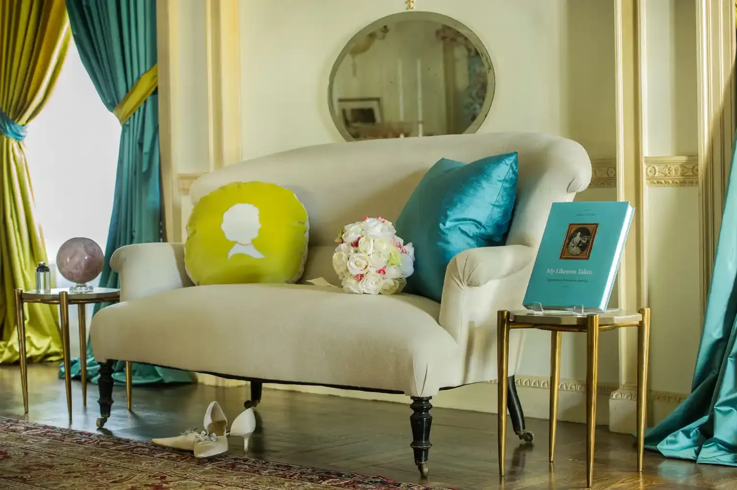 Elegant cream-colored sofa with teal and yellow pillows, a bouquet of white roses, a pair of white heels, and blue drapes in a luxurious room with a gold side table, a pink globe, and a turquoise book.