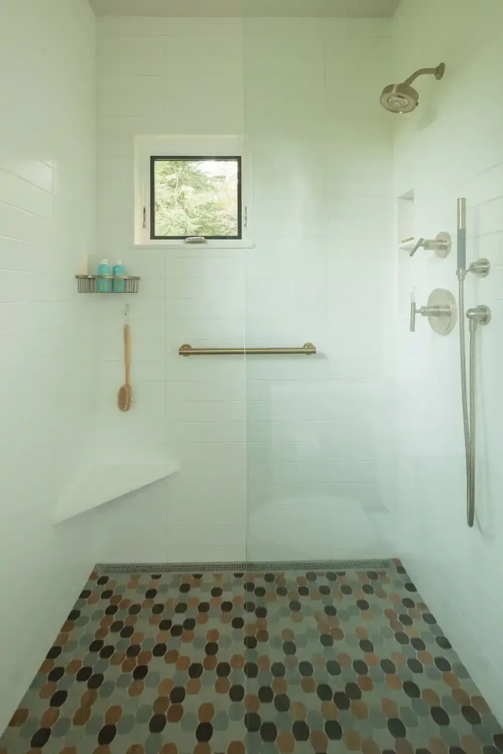 Empty walk-in shower with white tiled walls, a small window, a rain showerhead, a handheld shower, a wall-mounted shelf with toiletries, a bath brush, a grab bar, and a floor with multicolored hexagonal tiles.