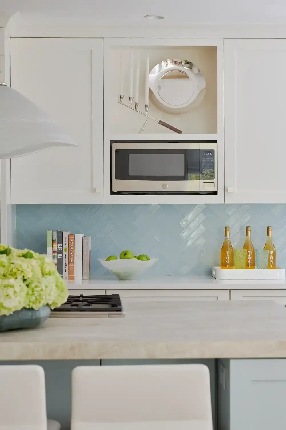 Modern kitchen with white cabinets, a microwave, a bowl of green apples, a row of books, and bottles of beverages on a tray.