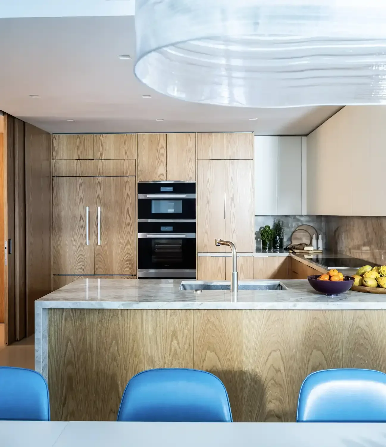 Modern kitchen with wooden cabinets, marble island, built-in oven, and a sink. Blue chairs are visible in the foreground.