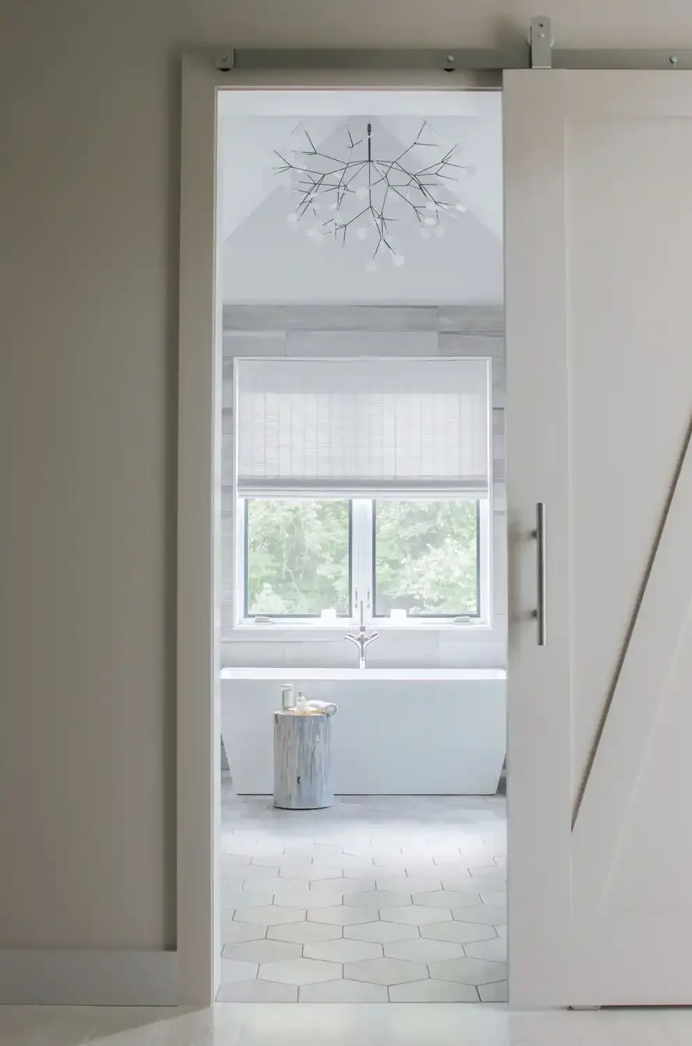 View through a partially open white sliding barn door into a modern bathroom with a bathtub, large window with blinds, and a small table made of wood.