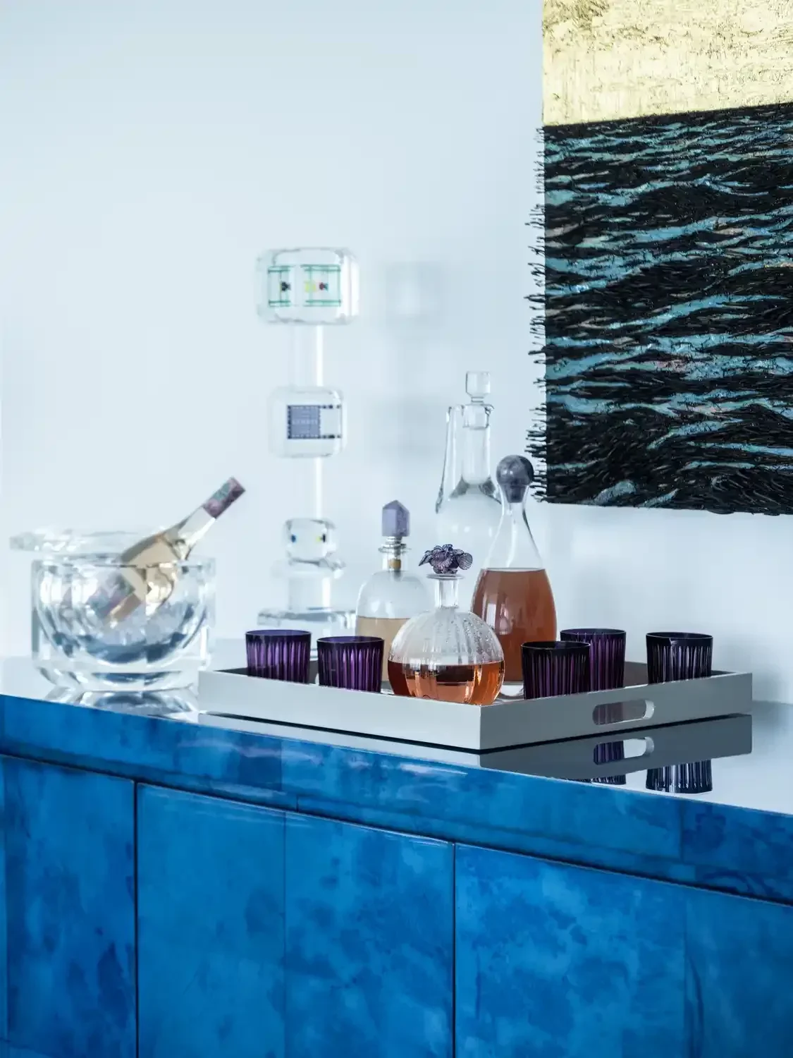 A blue cabinet with a white tray on top holding glass decanters and purple glasses; on the wall behind, there are modern glass fixtures and a textured black and beige painting.