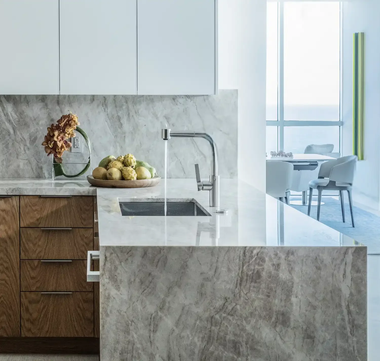 Modern kitchen with marble countertops, wooden cabinets, a bowl of fresh fruits, a vase with flowers, and a view of a bright dining area with large windows and white chairs.