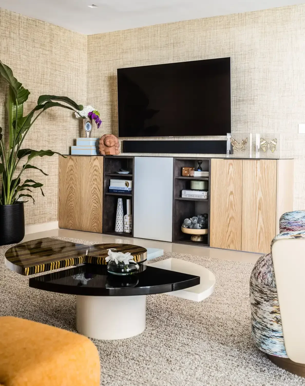 Living room with a wooden and dark cabinet, a large black TV mounted on the wall, a tall plant in a black pot, and a small round coffee table with a decorative tray. The room has textured beige wallpaper and a beige carpet.