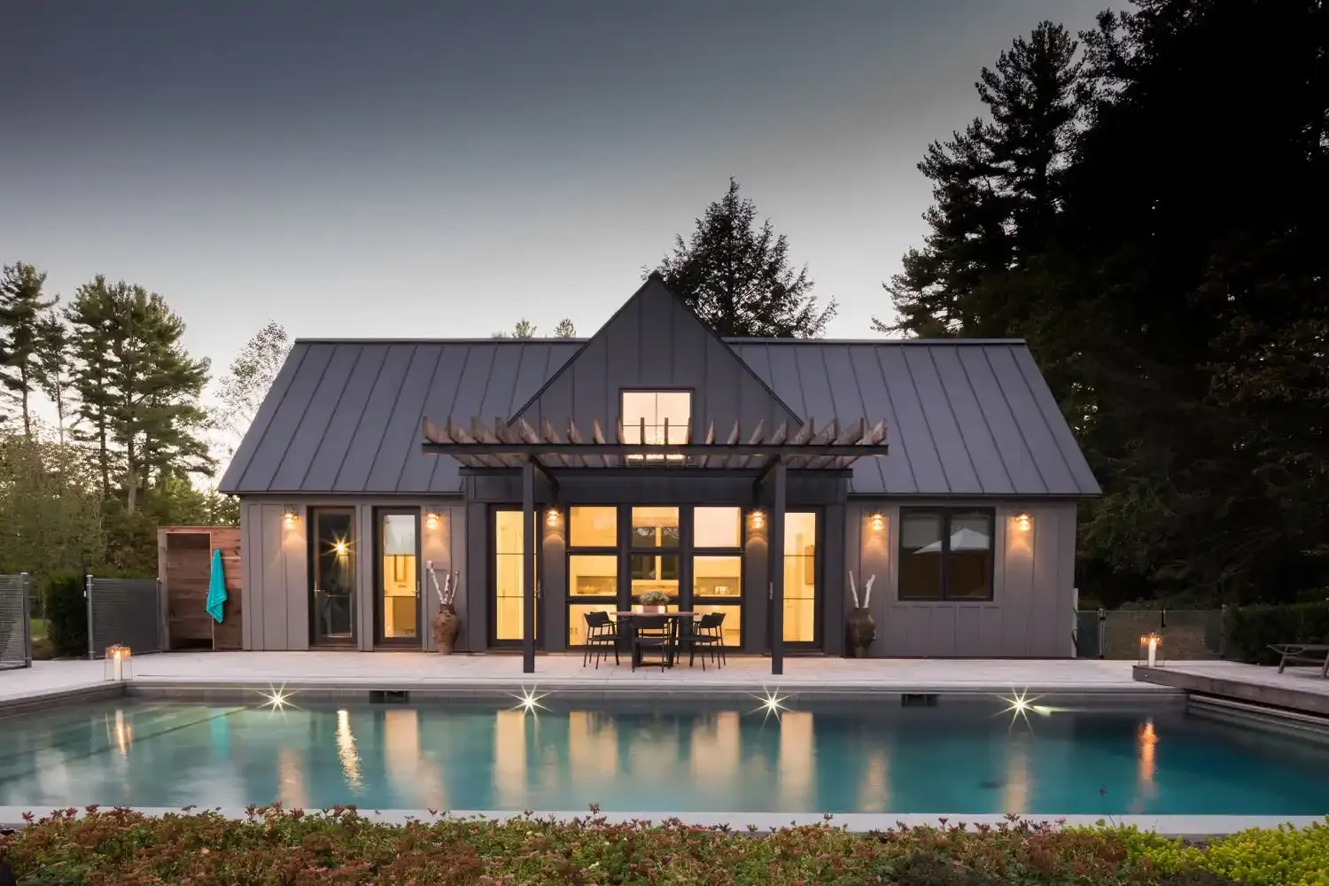 Modern house with black exterior, large windows, and a metal roof, situated behind a backyard swimming pool surrounded by a deck and greenery at dusk.