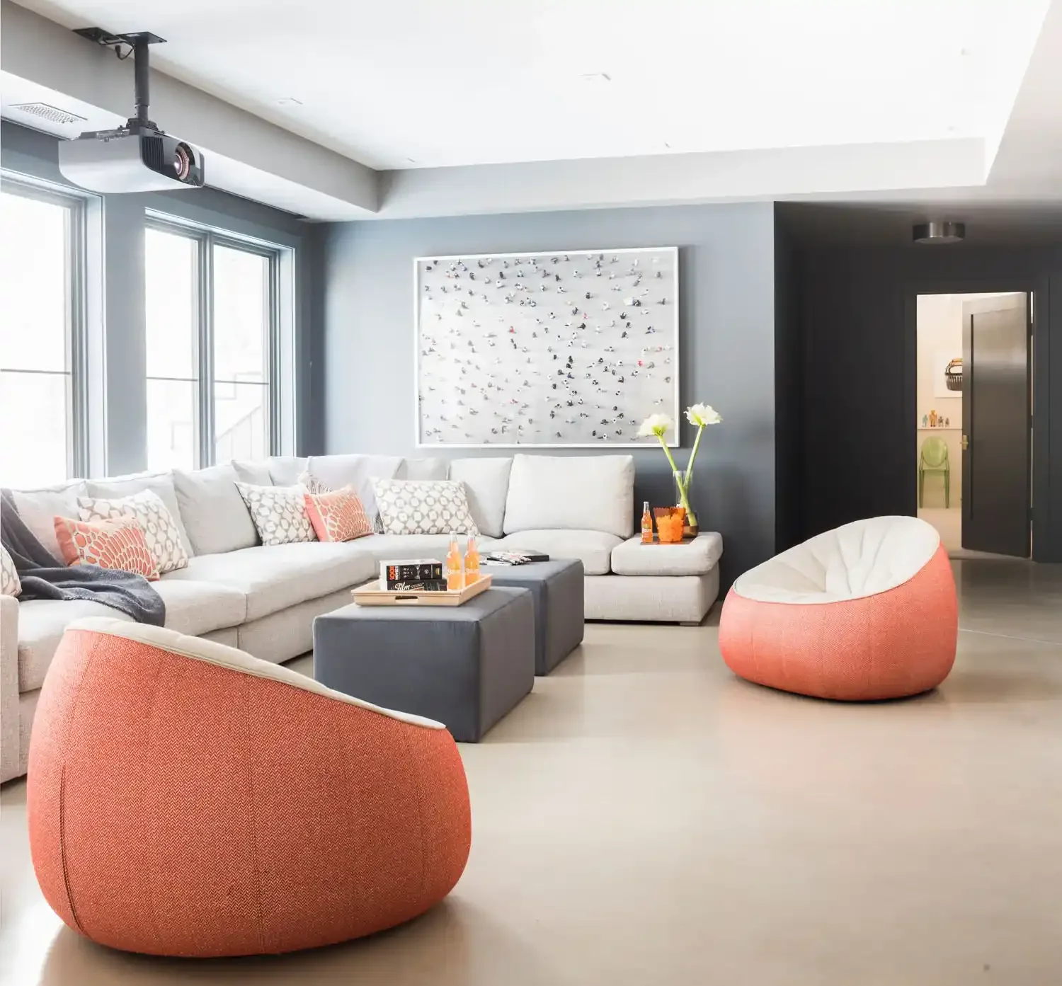 Modern living room with large white sectional sofa with patterned pillows, two orange chairs, and a dark gray ottoman. Decor includes a wall-mounted art piece, tall vases with flowers, and drinks on a tray. Large windows provide natural light, and an