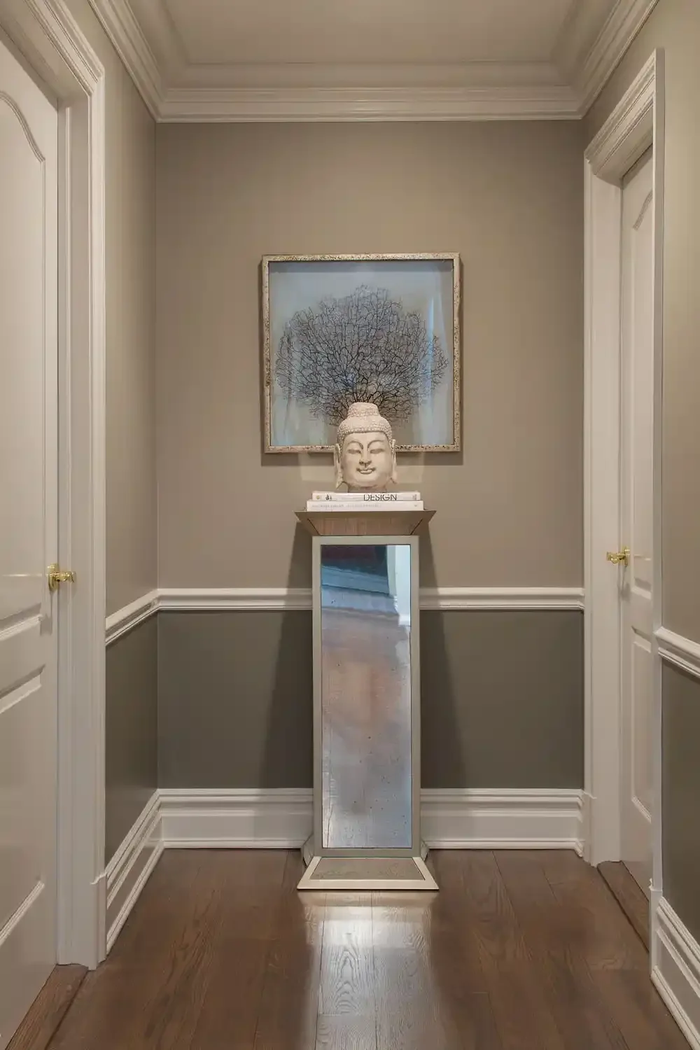 Hallway with white doors on each side, gray walls with white crown molding, hardwood floor, decorative sculpture of Buddha's head on a stand, and framed artwork of a coral branch behind the sculpture.