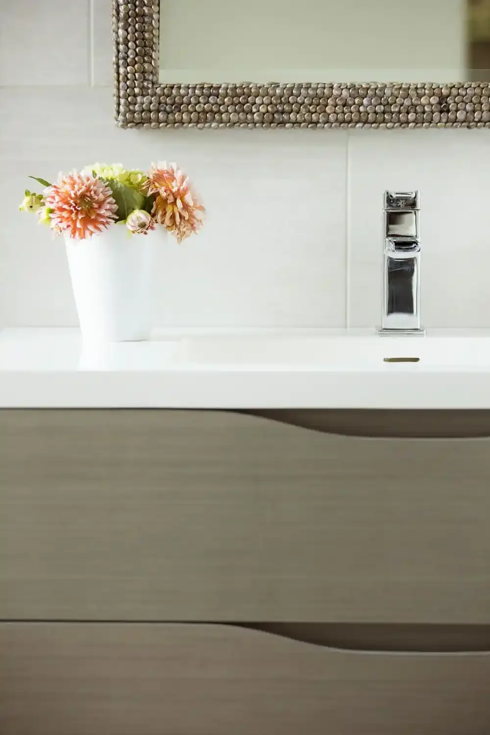 A white bathroom sink with a modern chrome faucet, a white vase with pink and green flowers, a mirror with a shell-like decorative frame, and a light-colored wooden cabinet below.