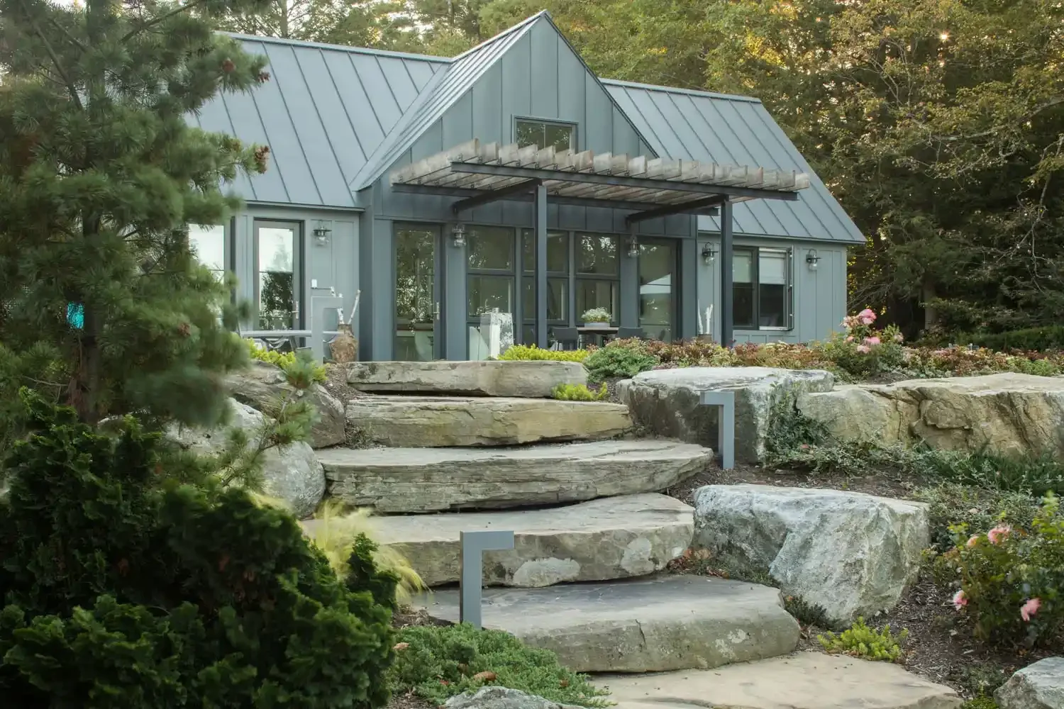 A modern gray house with a metal roof is situated on a landscaped yard with large rocks and plants, and stone steps leading to the entrance.