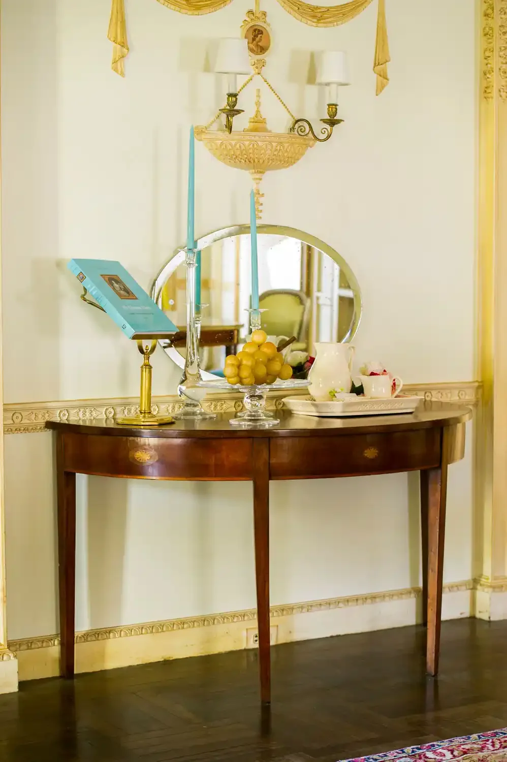 Wooden console table with a mirror, candles, a book, and a fruit bowl in a room with cream-colored walls and decorative trim.
