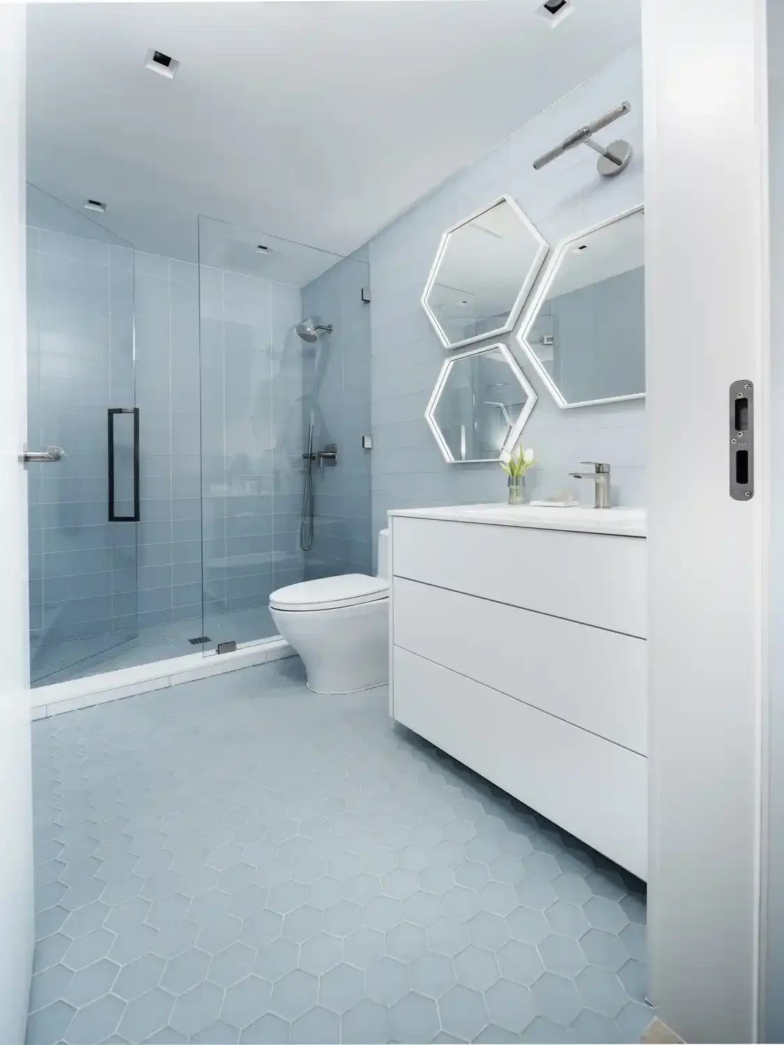 Modern bathroom with glass-enclosed shower, white toilet, and white vanity with hexagonal mirror frames and lighting.