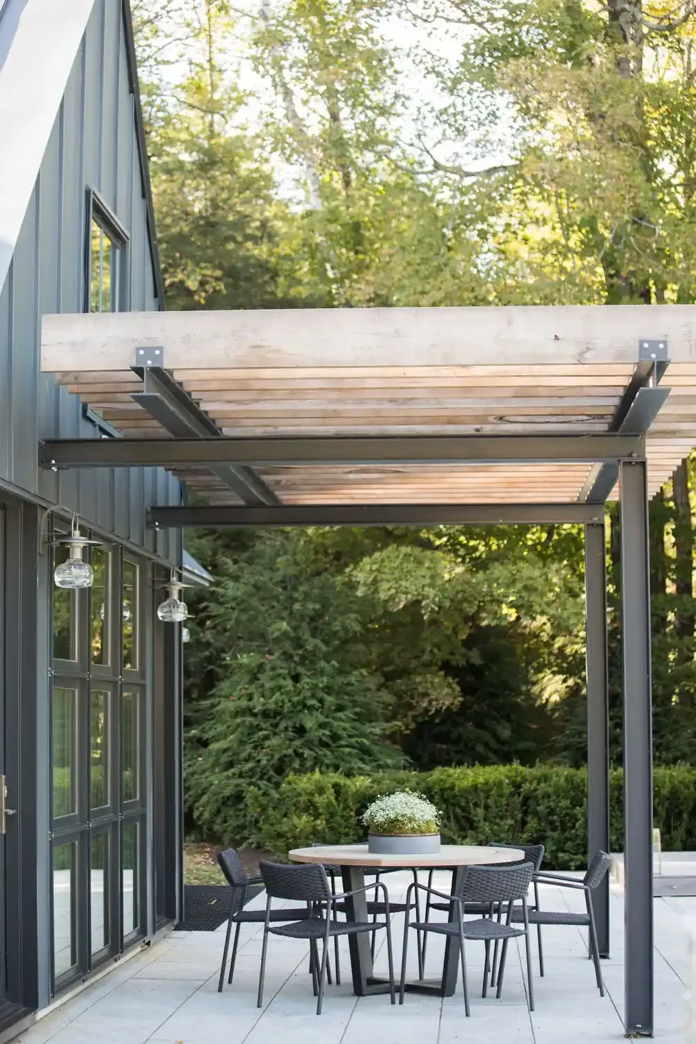 Outdoor patio with a round dining table, six chairs, and a potted plant in the center, under a wooden pergola on a concrete terrace, surrounded by greenery and a modern house with large glass doors.