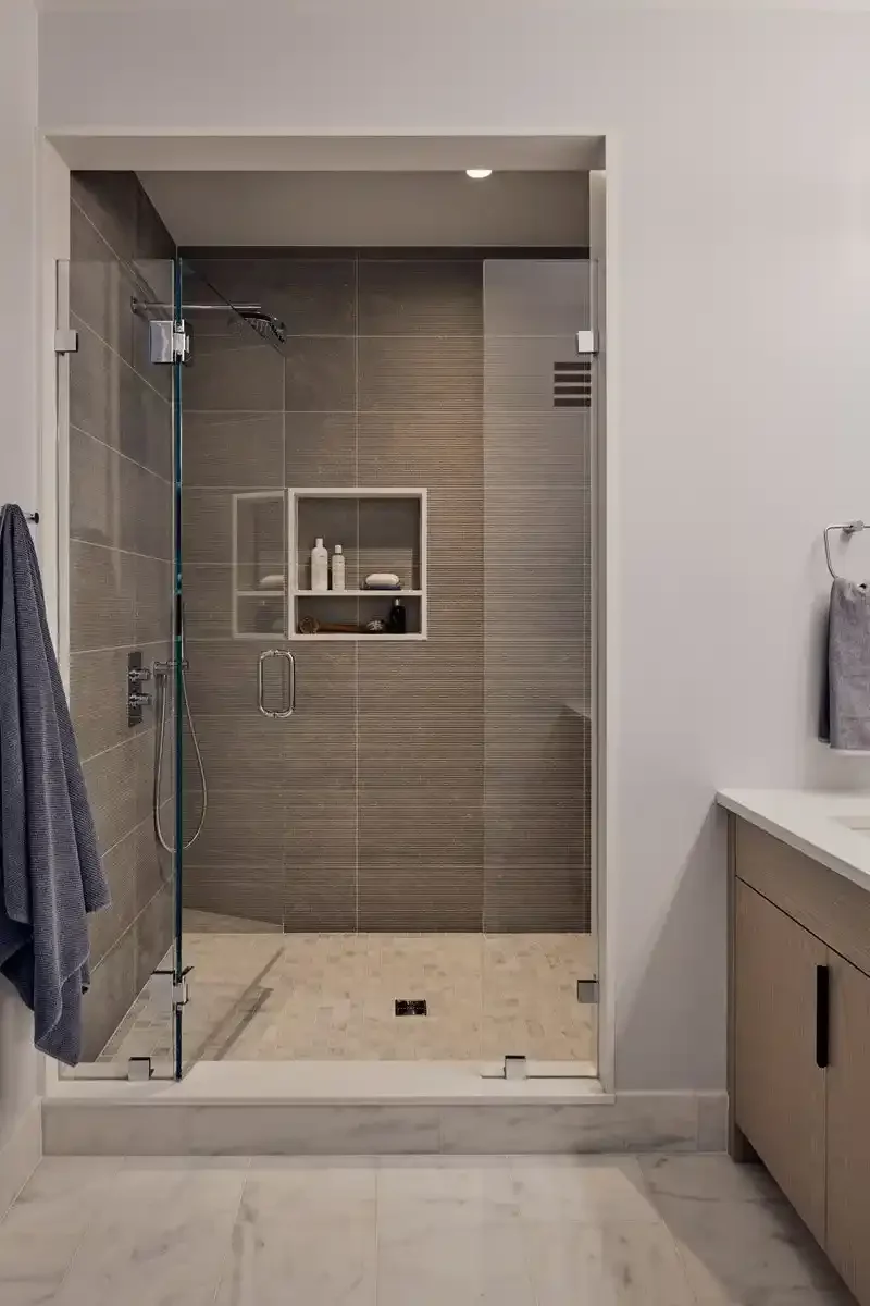 Modern glass-enclosed walk-in shower with built-in wall niche for toiletries, gray tiles, and a white vanity with a towel hanging on a hook.