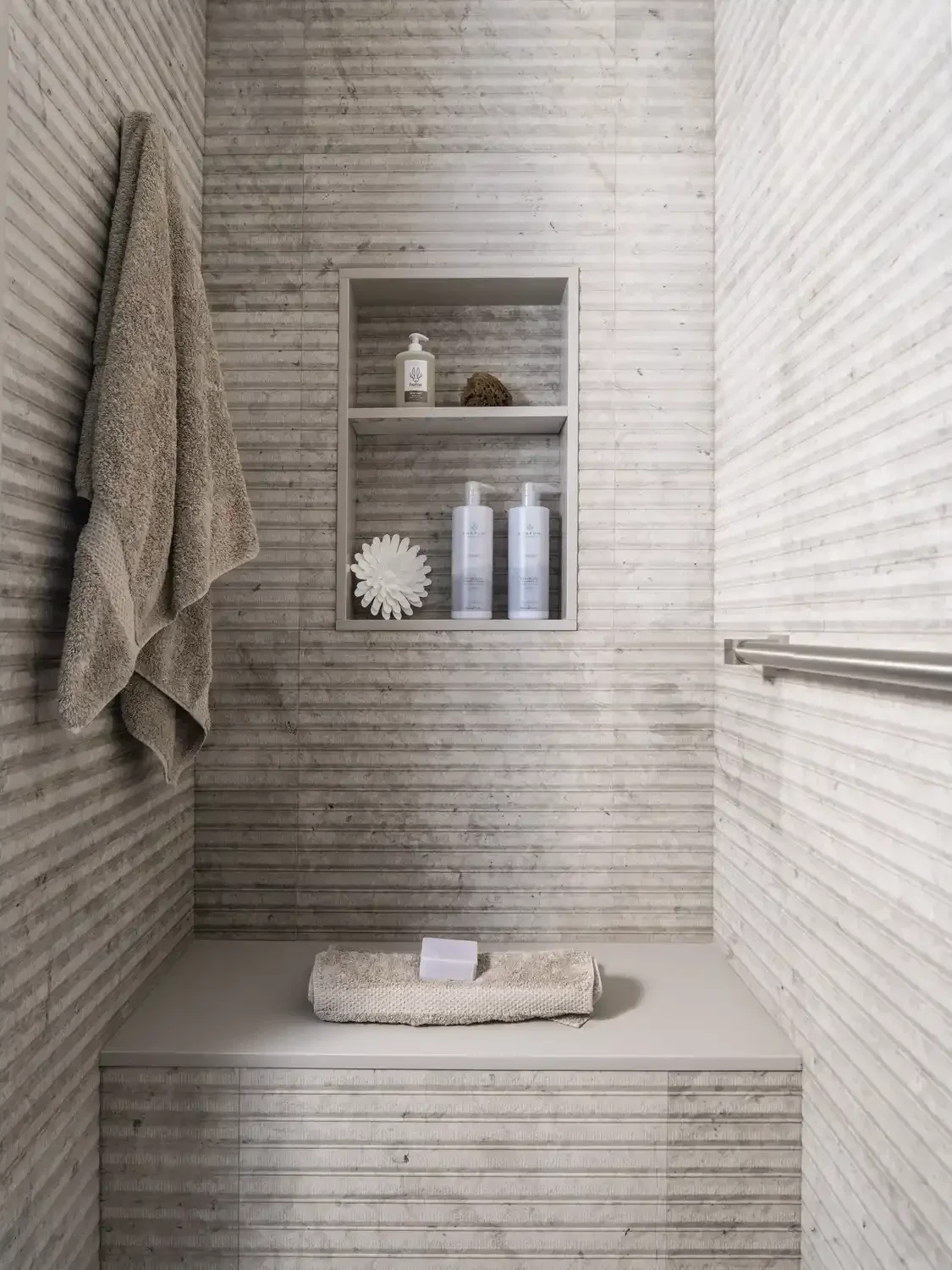 A minimalist shower with beige textured tiled walls, a built-in shelf holding shampoo bottles, a soap dispenser, a natural sponge, and decorative item, beige towels hanging on the side, a towel on the shower bench, and a horizontal metal grab bar on 